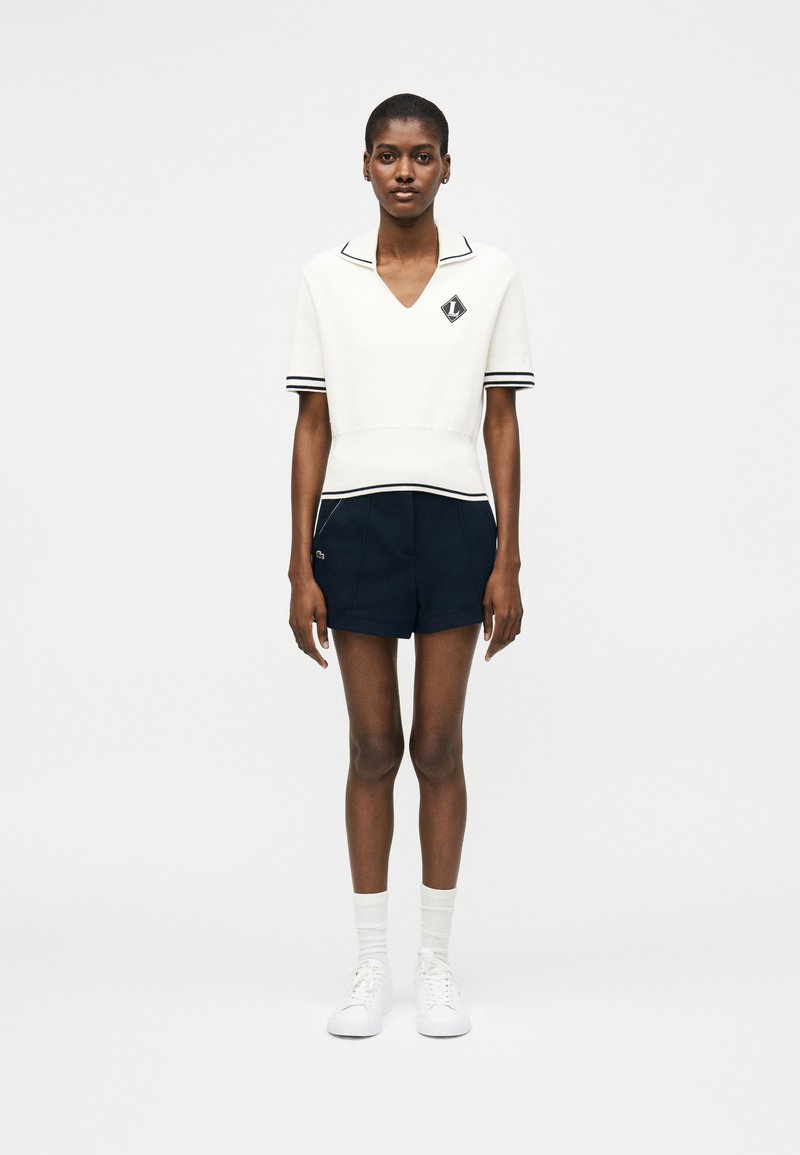 Femme debout, face à l'objectif, portant un pull blanc à manches courtes avec col en V, bordure noire et logo, un short marine, des chaussettes blanches et des baskets blanches.
