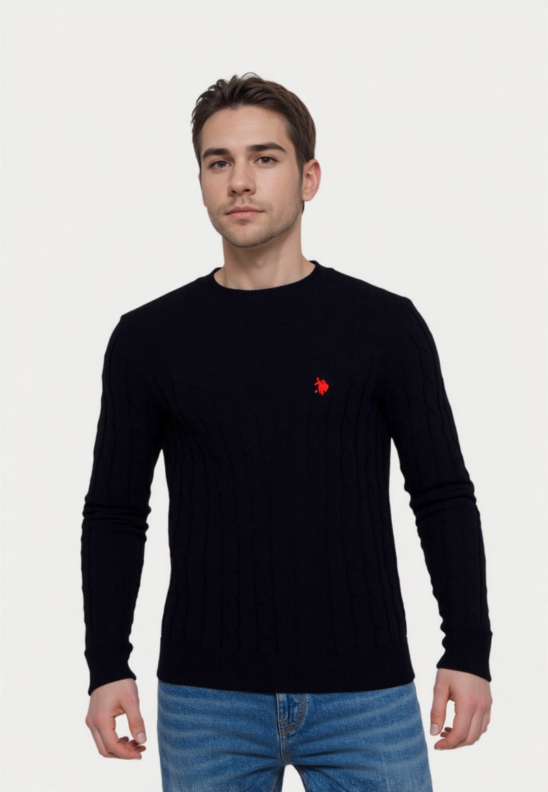 Tengerészkék, kötött pulóver hosszú ujjakkal és bordázott mandzsettákkal, bal oldalon kis piros logóval. Kék farmernadrággal viselve.