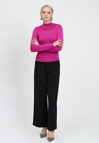 Donna in piedi con le braccia incrociate, che indossa un maglione di perline rosa brillante, pantaloni neri larghi e scarpe con tacco a disegni su sfondo bianco.