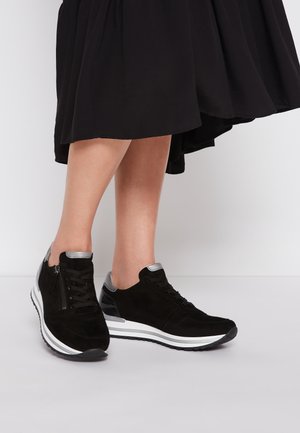 Persoon die zwarte sneakers met witte zolen en zilveren accenten draagt, gecombineerd met een zwarte rok, staand op een witte vloer.