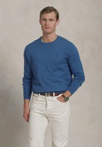 Blauwe gebreide trui met ronde hals, lange mouwen, gedragen met witte jeans en een bruine riem. Model staat casual met de handen in de zakken.