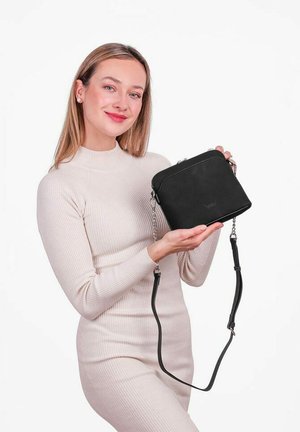 Woman in beige dress holds a small black crossbody bag with chain and leather strap against a plain white background.