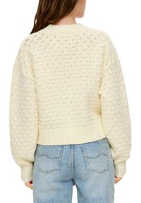Cremefarbener gestrickter Pullover mit Rautenmuster, geripptem Saum und puffärmel. Kombiniert mit hellblauen Jeans.
