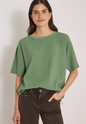 Femme portant un haut ample vert texturé à manches courtes et un pantalon marron foncé, debout avec une main dans la poche sur un fond uni.