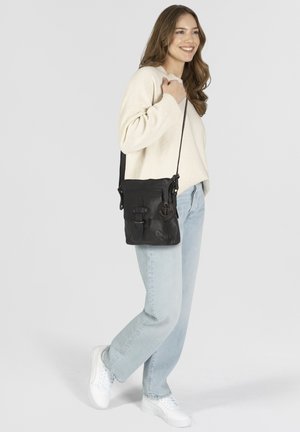Zwarte leren schoudertas met een gestructureerde afwerking, twee voorkantgespen en een metalen ankerbedel. Draagt bij lichte blauwe jeans en een crèmekleurige trui.