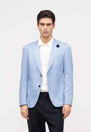 Ung mand iført en lyseblå blazer med mørke knapper, hvid skjorte, sorte bukser og en sort reversnål, stående foran en ensfarvet baggrund.