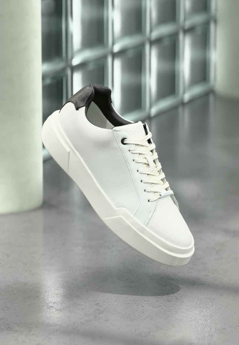Λευκό αθλητικό παπούτσι χαμηλού ύψους με μαύρη υφασμάτινη φτέρνα, λευκά κορδόνια και χοντρή σόλα, τοποθετημένο πάνω σε γυαλιστερό τσιμεντένιο πάτωμα κοντά σε γυάλινους τοίχους.