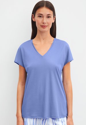 Frau mit glattem dunklem Haar, die ein hellblaues V-Ausschnitt-Top mit kurzen Ärmeln und gestreifte weiße und blaue Hose trägt, vor einem weißen Hintergrund steht.