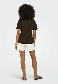 T-shirt di cotone marrone a maniche corte, abbinato a pantaloni corti bianchi con orlo sfrangiato. Il modello indossa sandali marroni.