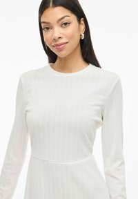 Robe blanche côtelée à manches longues avec un col rond, dotée d'un corsage ajusté et d'une jupe fluide. Texture de tissu lisse avec des lignes subtiles.