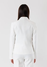 Blazer bianco su misura con rever a punta, linee pulite e maniche lunghe, caratterizzato da un tessuto liscio e strutturato e una silhouette aderente.