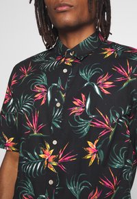 Korte mouw shirt, zwart stof met een levendig bloemdesign in roze, oranje en groen. Heeft een knoopsluiting kraag en tan knopen.