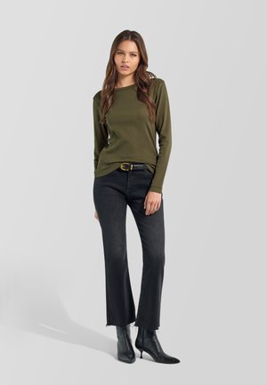 Donna con capelli castani lunghi che indossa una camicia a maniche lunghe color oliva, cintura nera, jeans neri e stivaletti neri con tacco, in piedi su sfondo grigio.