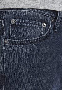 Vaqueros de denim azul oscuro con un diseño clásico de cinco bolsillos, acentos de remaches y una tela texturizada que muestra patrones de tejido visibles.