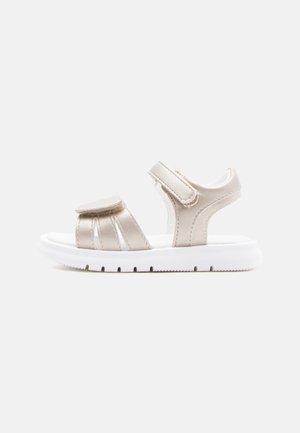 Beige sandal med åben tå, justerbare velcroremme og tyk hvid sål, set fra siden mod en ensfarvet baggrund.