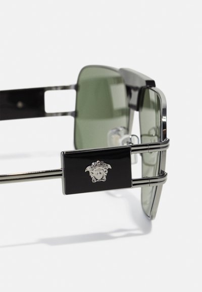 Lunettes de soleil avec lentilles vertes, monture en métal argenté, accents noirs et un fermoir décoratif mettant en avant un détail de logo au centre.