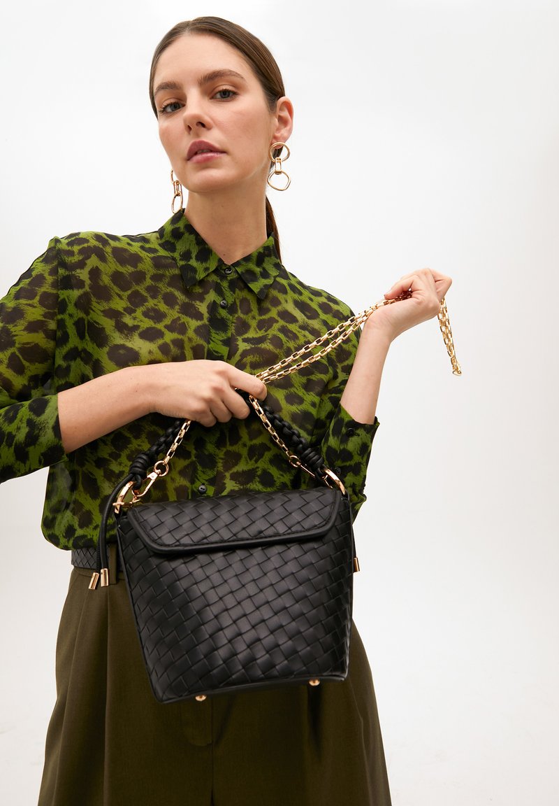 Svart vävd handväska med en slät yta, med ett flätat handtag, guldkedjeaxelrem och en strukturerad form. Grön djurmönstrad blus.