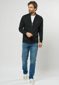 Sort zip-up jakke med høy krage og rødt logo, parret med blå denimjeans og lysegrå sneakers. Glatt, avslappet stoff.