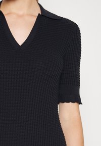 Černý pletený polo top s texturovaným vzorem, V-výstřihem, krátkými rukávy a vlnitými okraji na rukávových lemech.