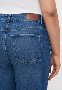 Denim jeans in dark blue with visible stitching, featuring a back pocket and a brown leather label on the waistband.