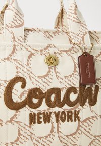 Beige canvas tas met bruin borduurd "Coach New York." Voorzien van een gouden drukknoopsluiting en een bruine leren label bevestigd met een ball chain.