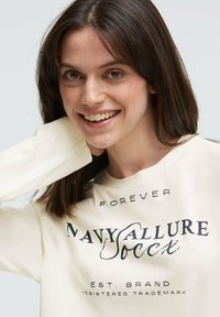 Langärmliges cremefarbenes Sweatshirt mit schwarzem Schriftzug: "FOREVER NAVY ALLURE", mit lockerem Schnitt und weicher Textur.