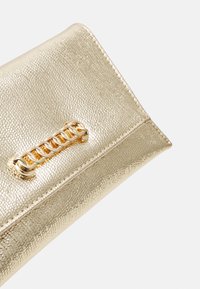 Pochette texturée dorée avec une courte chaîne dorée épaisse sur le rabat, posée sur un fond clair uni.