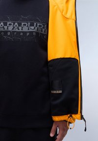 Černá mikina s texturovaným žlutým rukávem, s prasklým potiskem loga a elastickými detaily. Zapínání na zip a nastavitelné manžety.