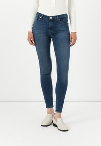 Jeans skinny en denim bleu foncé avec une texture lisse, design à cinq poches et fermeture à bouton. Associés à des chaussures slip-on de couleur claire.