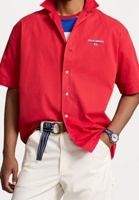 Red button-up shirt with short sleeves featuring a logo on the chest. Paired with white pants and a striped belt. Accessorized with a watch.