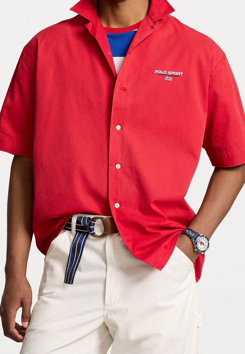 Red button-up shirt with short sleeves featuring a logo on the chest. Paired with white pants and a striped belt. Accessorized with a watch.