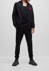 Černá mikina s kapucí na zip s malou červenou nášivkou loga, doplněná o černé tepláky ve stejném stylu. Nositel má také černé sportovní boty s barevnými akcenty.