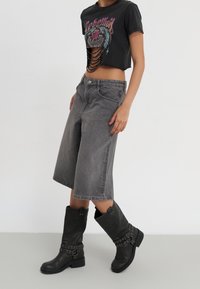 Camiseta negra recortada con diseño gráfico y detalles cortados. Pantalones cortos de denim anchos en color gris. Botas negras hasta la rodilla con acentos de hardware plateado.