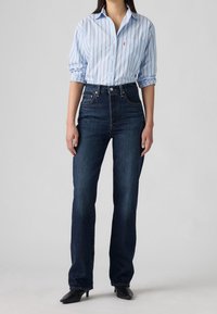 Kvinde iført en lyseblå skjorte med striber, knapper og stoppet ned i højtaljede, mørkeblå jeans med lige ben og sorte sko med hæl.