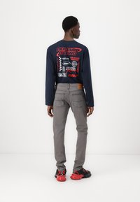 Marineblå langermet skjorte med Red Bull Racing-grafikk, grå denimbukser og sorte joggesko med røde detaljer, sett fra baksiden.