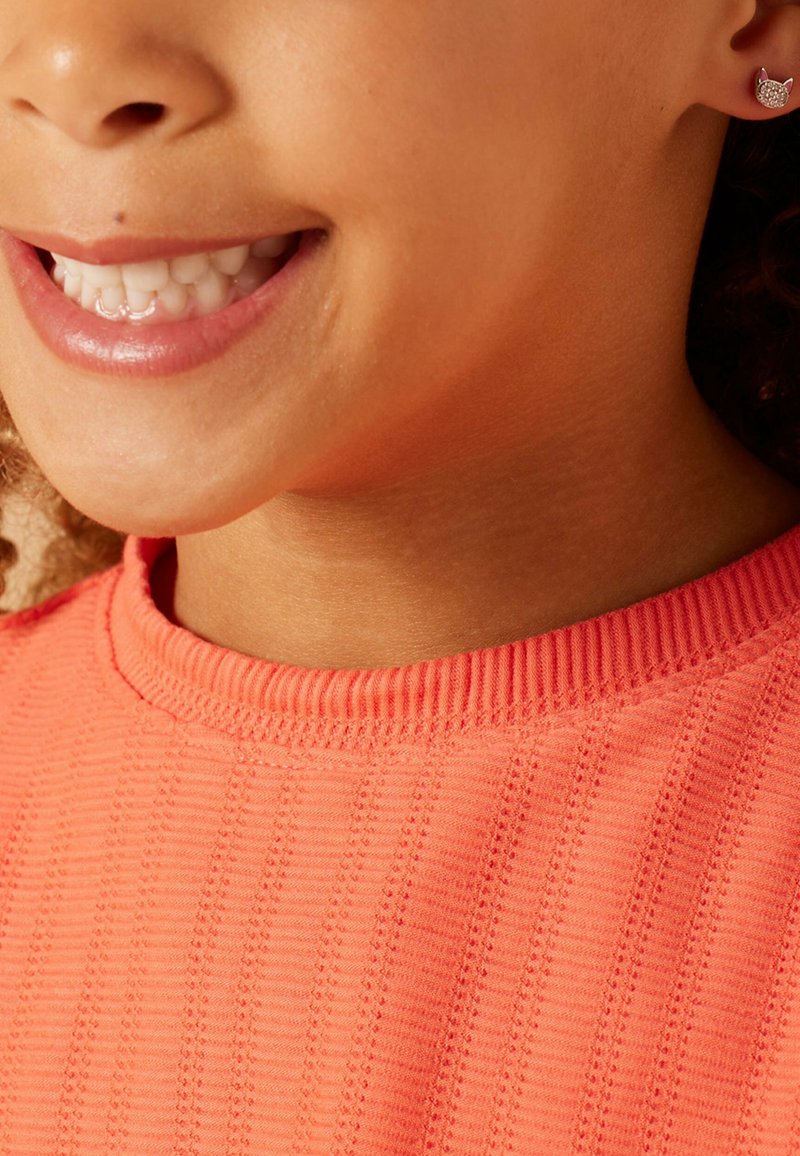 Koraalkleurig shirt met een geribbeld halslijn, gedragen door een kind, met kleine kattenvormige studs met een glinsterende oppervlakte.
