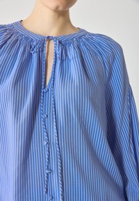 Blåstripet bluse med rynket halslinje, knappeåpning og flytende ermer. Glatt stoff med vertikale hvite striper.