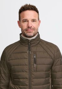 Mann med kort brunt hår og skjeggstubber iført en olivengrønn quiltet jakke over en beige genser med rullekrage, som vender mot kameraet foran en ensfarget bakgrunn.