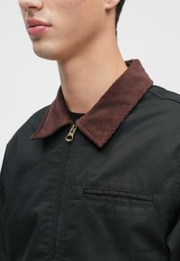 Artimas žmogaus, vilkinčio juodą striukę su rudomis veliūrinėmis apykaklėmis ir auksiniu užtrauktuku, vaizdas, matomas dalis veido ir kaklo.