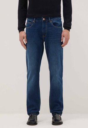 Homme portant un jean droit bleu, une chemise noire à manches longues et des mocassins en cuir noir, debout devant un fond uni.