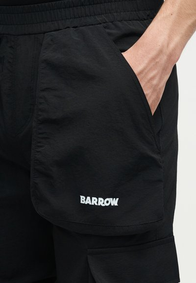 Χέρι στην τσέπη μαύρου σορτς με ελαστική μέση και μεγάλη πλαϊνή τσέπη που φέρει λευκό λογότυπο "BARROW".
