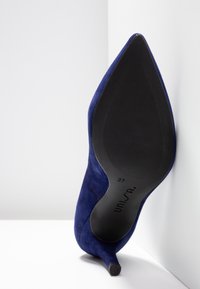 Chaussure à talon haut en daim bleu marine à bout pointu avec semelle noire, taille 37, positionnée verticalement contre une surface d'angle blanche.