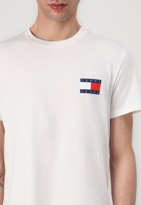 Biely bavlnený tričko s krátkymi rukávmi, vybavené malým obdĺžnikovým logom v modrej, červenej a bielej farbe na ľavej oblasti hrude.