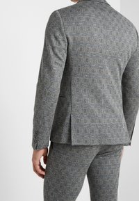 Grijze op maat gemaakte blazer met een gestructureerd ruitpatroon, voorzien van een enkele split aan de achterkant en knopen op de manchetten.