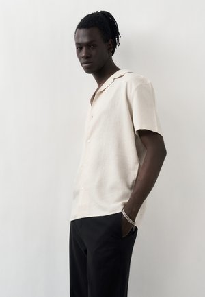 Młody mężczyzna o ciemnej karnacji i dredach, ubrany w kremową koszulę z krótkim rękawem na guziki i czarne spodnie, stojący na jednolitym białym tle.