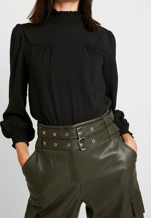 Femme portant un chemisier noir à manches longues avec col et poignets volantés, associé à un pantalon en cuir vert olive taille haute avec double boucles de ceinture.