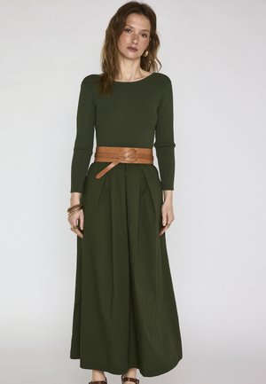 Donna con lunghi capelli castani che indossa una maglia a maniche lunghe verde scuro e pantaloni ampi abbinati, accentuati da una cintura avvolgente marrone e gioielli dorati.