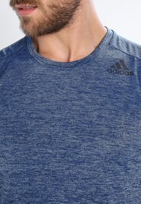 Bearded man wearing a blue heathered Adidas t-shirt, visible from the neck to mid-chest, looking to the left.