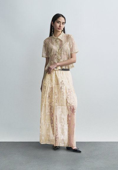 Γυναίκα με μπεζ διαφανές δαντελένιο πουκάμισο και μακριά φούστα με λουλουδάτα σχέδια, στέκεται σε γκρι πάτωμα μπροστά σε λευκό απλό τοίχο.