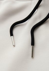 Biała tkanina z czarnymi plecionymi sznurkami, zakończonymi metalicznymi srebrnymi końcówkami. Materiał wydaje się miękki i gładki.